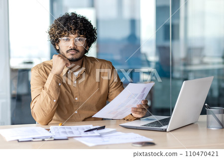 Portrait of serious concentrated businessman inside office, man looking at camera holding papers financial reports, accountant financier with laptop on paper work. 110470421