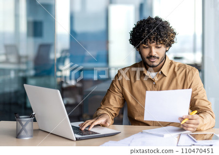 A young businessman with curly hair sits in an office, immersed in his laptop, crafting a digital masterpiece. 110470451