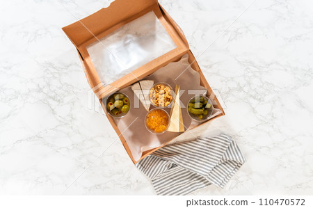 Flat lay. In the process of assembling, a woman expertly curates this charcuterie box, showcasing sliced meat, cheese, crackers, and grapes, all neatly arranged and packaged in a brown gifting box. 110470572