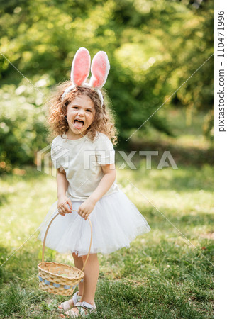 Easter egg hunt. Girl child Wearing Bunny Ears Running To Pick Up Egg In Garden. Easter tradition. Baby with basket full of colorful eggs. Easter egg hunt. Girl child Wearing Bunny Ears Running To Pick Up Egg In Garden. Easter tradition. Baby with basket full of colorful eggs. 110471996