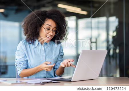 Businesswoman at the workplace uses a laptop for a video call, the employee happily smiles and friendly talks with colleagues and partner at an online meeting, works inside the office. 110472224