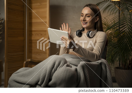Video conference. Portrait of a cheerful woman in a headset making a web call using a tablet computer, sitting on an upholstered chair, talking, waving at a webcam Video conference. Portrait of a cheerful woman in a headset making a web call using a tablet computer, sitting on an upholstered chair, talking, waving at a webcam 110473389