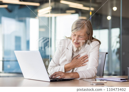 Heart attack chest pain, senior mature businesswoman in business suit working inside office at workplace with laptop, having severe heart pain holding hand to chest. 110473402
