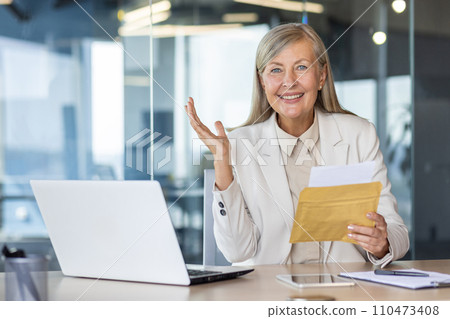 Senior mature business woman boss inside office at workplace, woman smiling joyfully and looking at camera, holding mail envelope notification letter notification of successful achievement results. Senior mature business woman boss inside office at workplace, woman smiling joyfully and looking at camera, holding mail envelope notification letter notification of successful achievement results. 110473408