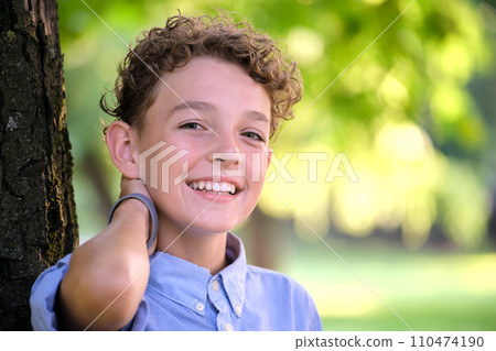 Young happy child boy relaxing in summer park leaning on tree trunk. Child wellbeing concept 110474190