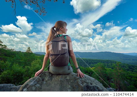 Sportive woman sitting alone taking a break on hillside trail. Female hiker enjoying view of nature from rocky cliff on wilderness path. Active lifestyle concept 110474414
