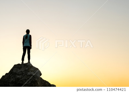Silhouette of a woman hiker standing alone on big stone at sunset in mountains. Female tourist on high rock in evening nature. Tourism, traveling and healthy lifestyle concept. Silhouette of a woman hiker standing alone on big stone at sunset in mountains. Female tourist on high rock in evening nature. Tourism, traveling and healthy lifestyle concept. 110474421