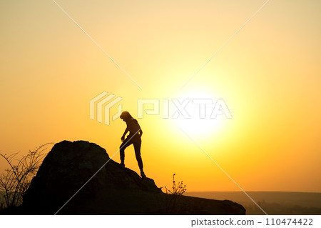 Silhouette of a woman hiker climbing up a big stone at sunset in mountains. Female tourist on high rock in evening nature. Tourism, traveling and healthy lifestyle concept. Silhouette of a woman hiker climbing up a big stone at sunset in mountains. Female tourist on high rock in evening nature. Tourism, traveling and healthy lifestyle concept. 110474422
