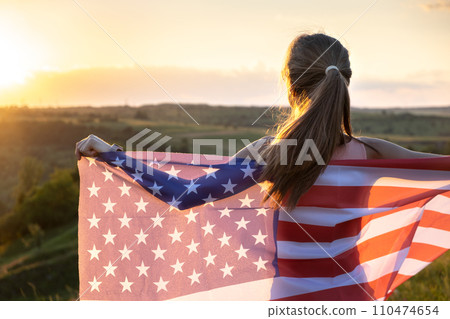 Happy young woman posing with USA national flag standing outdoors at sunset. Positive girl celebrating United States independence day. International day of democracy concept. 110474654