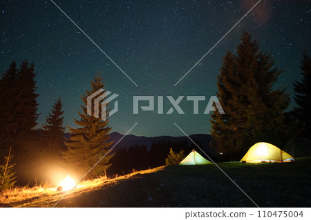 Bright illuminated tourist tents near glowing bonfire on camping site in dark mountains under night sky with sparkling stars. Active lifestyle concept. 110475004