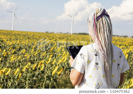 A young woman with a tablet in a field with sunflowers, wind turbines for green energy production, eco-energy, wind turbines A young woman with a tablet in a field with sunflowers, wind turbines for green energy production, eco-energy, wind turbines 110475362