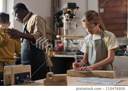 Waist up portrait of blonde young girl measuring wooden board in carpenting workshop for children Waist up portrait of blonde young girl measuring wooden board in carpenting workshop for children 110476808