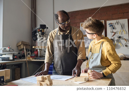 Portrait of Black senior carpenter teaching little boy woodworking in workshop and looking at plans Portrait of Black senior carpenter teaching little boy woodworking in workshop and looking at plans 110476832