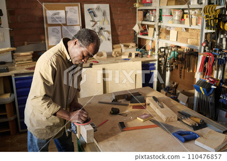 High angle shot of male carpenter building wooden furniture in sunlit workshop and polishing planks High angle shot of male carpenter building wooden furniture in sunlit workshop and polishing planks 110476857