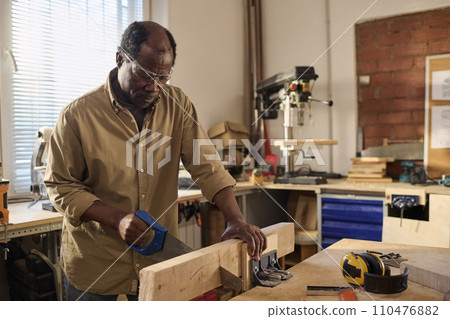 Portrait of African American senior man cutting wood while building furniture in sunlit workshop 110476882