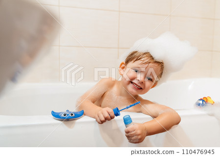 Smiling boy with a funny foam hat on his head, taking a bath with foam, toys and bubbles. 110476945