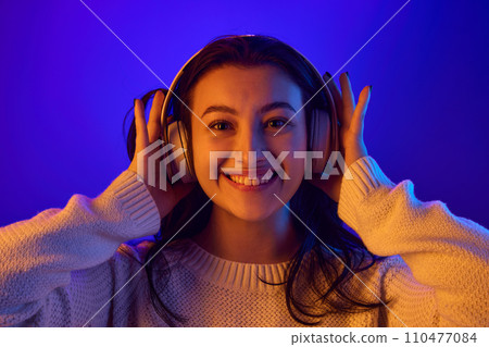 Portrait of young attractive woman puts on fashionable modern headphones and smiling looking at camera against gradient blue background. 110477084