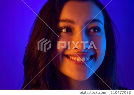 Close up portrait of young attractive woman smiling and looking away against gradient blue background in neon light. Close up portrait of young attractive woman smiling and looking away against gradient blue background in neon light. 110477088