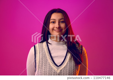 Young woman with long brunette hair, dressed casual outfit smiling looking at camera against gradient pink background. 110477146