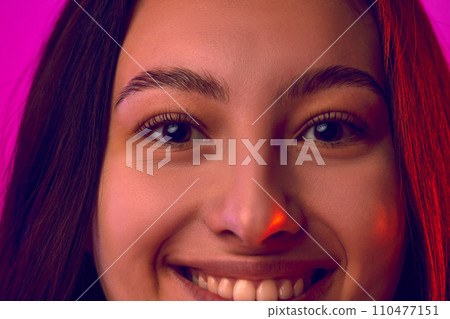 Extremely close up cropped portrait of face of young attractive woman smiling looking to camera against gradient pink background. 110477151