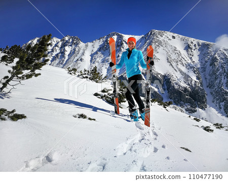 Young woman in light blue jacket standing, smiling happy holding with mountaineering ski and poles in hands, sun shines on snow, bright sky over mountains behind 110477190