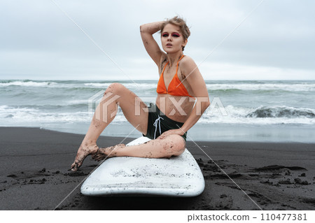 Woman surfer sitting on surfboard on sandy beach on background of ocean waves during summer holiday 110477381