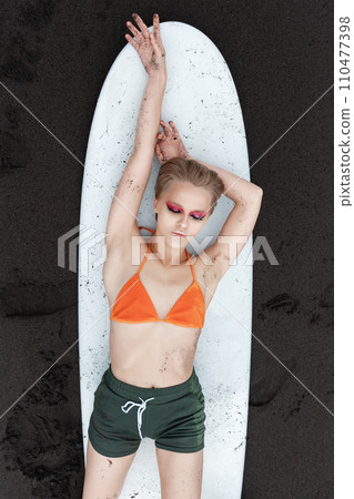 Top view slim female surfer looks so relaxed and at peace, lying on surfboard on black sandy beach Top view slim female surfer looks so relaxed and at peace, lying on surfboard on black sandy beach 110477398