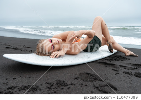 Female surfer lying on white surfboard on black sandy beach. Sports fashion model with closed eyes 110477399