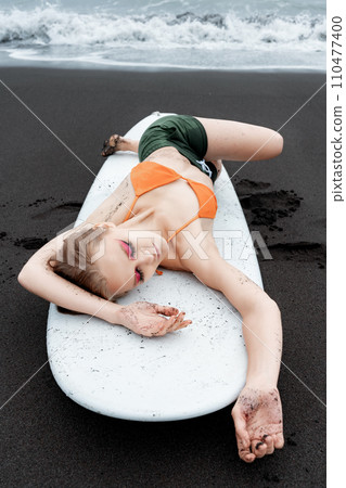 Female surfer is enjoying well deserved break on sandy beach, lying on surfboard with eyes closed 110477400