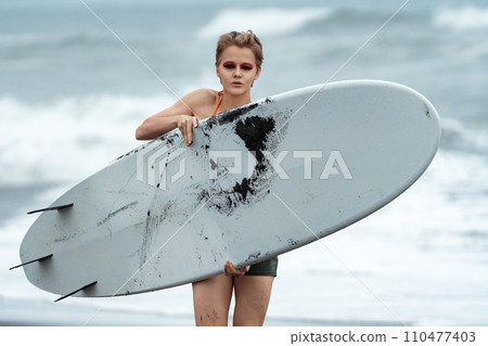 Woman surfer carrying white surfboard walking on beach, looking at camera on background of sea waves 110477403