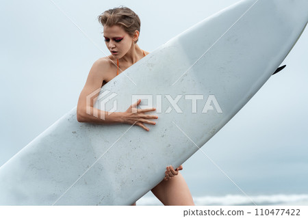 Close-up view of female sports fashion model holding white surfboard and standing behind it Close-up view of female sports fashion model holding white surfboard and standing behind it 110477422