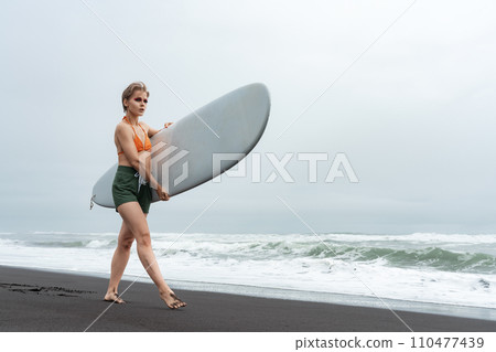 Female surfer walks along black sand beach while carrying surfboard against backdrop of ocean waves 110477439