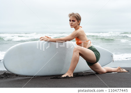 Female surfer is standing on knee, stretching one leg forward, and holding surfboard with one hand Female surfer is standing on knee, stretching one leg forward, and holding surfboard with one hand 110477445