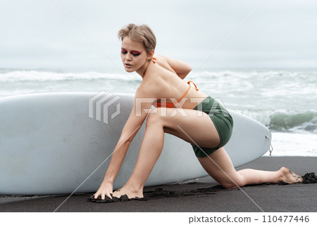Female surfer stands on one knee, stretching leg forward on beach, and surfboard with one hand Female surfer stands on one knee, stretching leg forward on beach, and surfboard with one hand 110477446