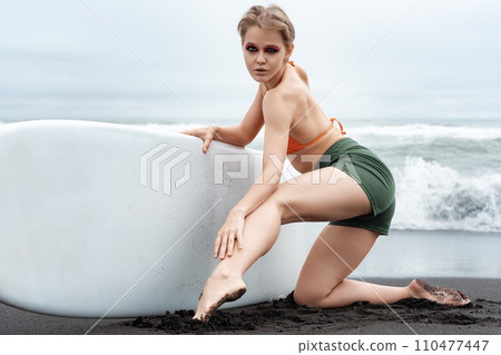 Female surfer exudes confidence as she kneels on black sand beach, holding surfboard with ease Female surfer exudes confidence as she kneels on black sand beach, holding surfboard with ease 110477447