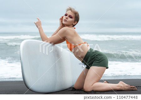 Female surfer exudes confidence as she gracefully kneels on black sand beach, holding surfboard Female surfer exudes confidence as she gracefully kneels on black sand beach, holding surfboard 110477448