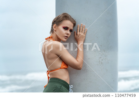 Woman holding surfboard vertically and pressing herself against it in sexual manner, looking away Woman holding surfboard vertically and pressing herself against it in sexual manner, looking away 110477459