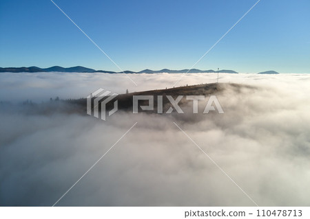 Aerial view of colorful sunrise over white dense fog with distant dark silhouettes of mountain hills on horizon Aerial view of colorful sunrise over white dense fog with distant dark silhouettes of mountain hills on horizon 110478713