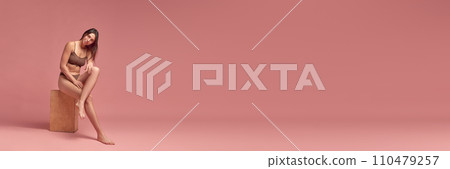 Beauty and wellness blog. Beautiful young girl with slim, fit body sitting, posing in cotton underwear against pink studio background. Beauty and wellness blog. Beautiful young girl with slim, fit body sitting, posing in cotton underwear against pink studio background. 110479257