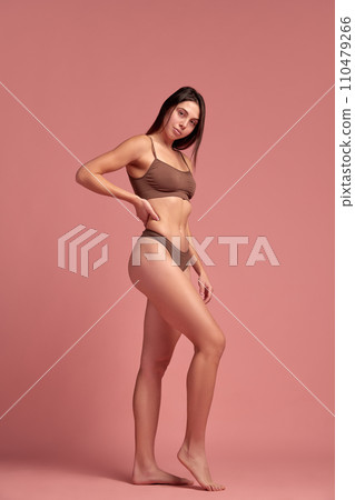 Loosing weight in healthy way. Young beautiful woman with slim body standing in cotton underwear against pink studio background. 110479266