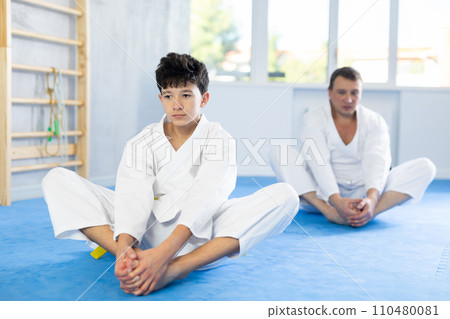 Teenage boy and middle-aged man trainer wearing kimono doing leg stretching in sports hall 110480081