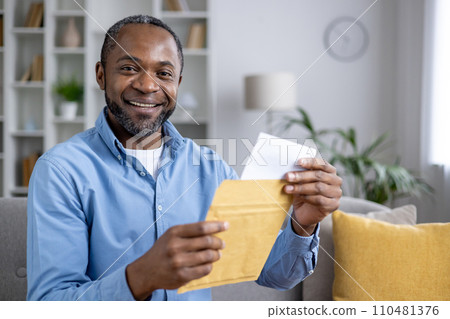 Close-up portrait of a smiling African-American man sitting at home and holding an open envelope with a letter in his hands, looking and smiling at the camera. 110481376