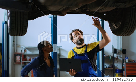 Mechanic in auto repair shop standing with customer underneath suspended car, looking together for replacement parts. Employee helps woman fix vehicle, using laptop to find components Mechanic in auto repair shop standing with customer underneath suspended car, looking together for replacement parts. Employee helps woman fix vehicle, using laptop to find components 110481750
