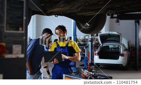 Trained mechanic and customer standing underneath car in garage using laptop to order new parts after finding defects. Employee assisting african american client by looking for components online Trained mechanic and customer standing underneath car in garage using laptop to order new parts after finding defects. Employee assisting african american client by looking for components online 110481754