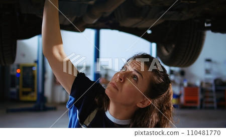 Qualified engineer working on suspended car in garage, checking components during routine maintenance. Auto repair shop employee underneath vehicle using work light to look for potential issues 110481770