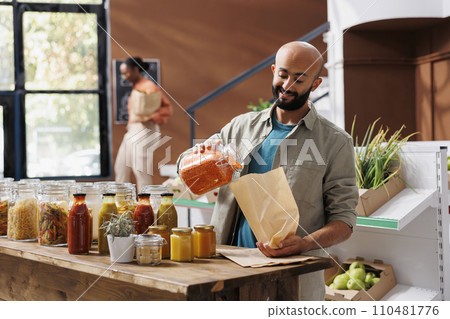 Smiling man pouring orange lentils into bag, buying healthy goods from reusable glass jars with organic bulk products. Customer purchasing natural bio food items from local eco zero waste store. 110481776