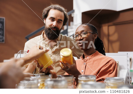 Close-up of multicultural couple in a plastic free convenience store, looking at glass jars of honey. African American girlfriend and her Caucasian boyfriend are shopping sustainably at local shop. Close-up of multicultural couple in a plastic free convenience store, looking at glass jars of honey. African American girlfriend and her Caucasian boyfriend are shopping sustainably at local shop. 110481826
