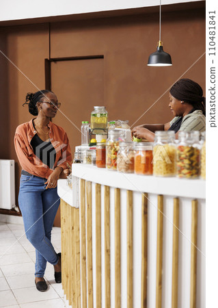 African American vendor checks out groceries at a local market offering eco-friendly, organic products. Black women at cashier desk conversing about locally grown nutritious organic products. African American vendor checks out groceries at a local market offering eco-friendly, organic products. Black women at cashier desk conversing about locally grown nutritious organic products. 110481841