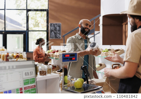 In a local zero waste eco store, a merchant scans and weighs goods to sell to a frequent customer. Caucasian vendor weighs fruits and vegetables and sells locally grown produce. In a local zero waste eco store, a merchant scans and weighs goods to sell to a frequent customer. Caucasian vendor weighs fruits and vegetables and sells locally grown produce. 110481842