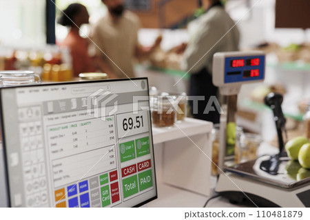 While multiethnic clients are assisted by salesman in the background, apples are placed on digital measuring scale. Photo focus on cashier's desk with weighing equipment and desktop computer. 110481879